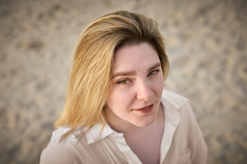 Young woman with blond hair gazes softly towards camera, standing on sandy beach. Her white shirt...