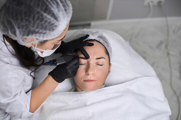Professional performs eyebrow permanent makeup procedure on relaxed adult woman in clean, tranquil...
