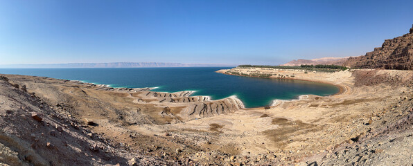 Panoramic view of the Dead Sea shoreline in Jordan with salt patterns, turquoise water, and desert terrain under a clear blue sky.