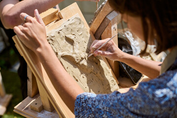 Young adult female sculpting clay relief portrait on wooden easel. Sunlight illuminates area,...