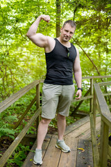 Confident muscular man flexing bicep in a sleeveless shirt and shorts while standing on a wooden bridge in a lush green forest. Hiking, fitness, lifestyle, outdoor activity, or motivation concepts