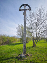 Wooden wayside cross with Jesus Christ crucifix standing in a green meadow near a path, under a spring sky.