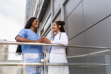 Multiracial female Doctor and nurse having a coffee break outside the hospital, smiling and talking