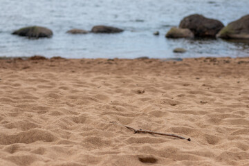 A single stick lies on textured sand near a rocky shoreline, with calm sea water softly blurred in...