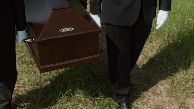 Low section of unrecognizable pallbearers wearing black suits and white gloves carrying elegant wooden casket along cemetery during funeral ceremony