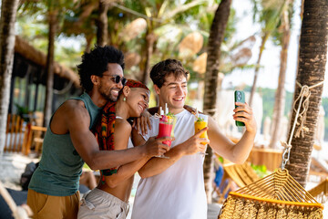 Group of cheerful young friends taking a selfie while holding colorful tropical drinks at a beach resort, capturing joyful summer memories
