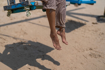 Bare feet dangle from a wooden swing above the sandy beach, casting soft shadows under the sun, capturing a carefree and peaceful summer moment.