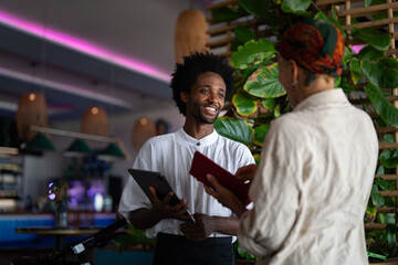 A smiling waiter is taking an order from a customer at a stylish restaurant, holding a tablet while engaging in friendly conversation.