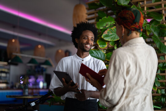 A smiling waiter is taking an order from a customer at a stylish restaurant, holding a tablet while engaging in friendly conversation.