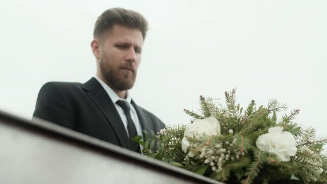 Young Caucasian man in black suit looking at casket at outdoor funeral and crying, grieving loss of loved one