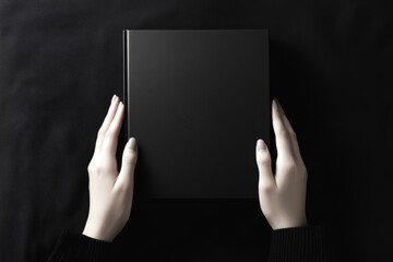 Black hardcover book held by two hands on black textured background. Top view close-up photography with minimalistic style. Book mockup and monochrome concept for design and print.