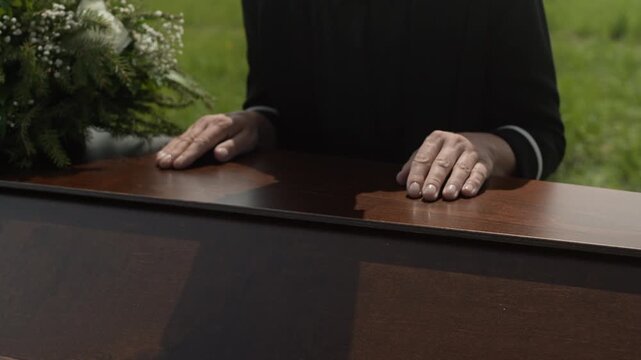 Slowmo of grieving widow in black clothes touching gently wooden coffin lid and then bowing her head feeling pain of loss at outdoor funeral ceremony