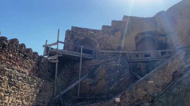 Camera clip tilts up weathered stone walls, revealing temporary scaffolds against frescoed cliffs under harsh noon light. Ideal for heritage conservation stories.