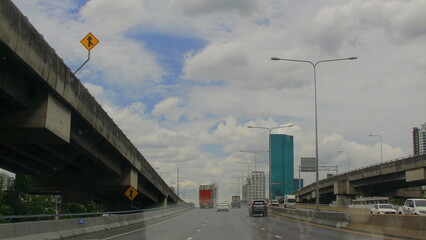 A multi-lane elevated highway curving into central Bangkok, with high-rises in the background and streetlights lining the route under a cloudy sky.