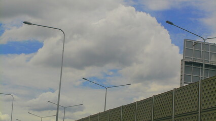 A view of a Bangkok highway beneath a dramatic cloudy sky, lined with tall lampposts and noise barriers, reflecting urban travel infrastructure.