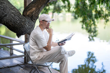man is sitting on a chair by a lake, reading a book
