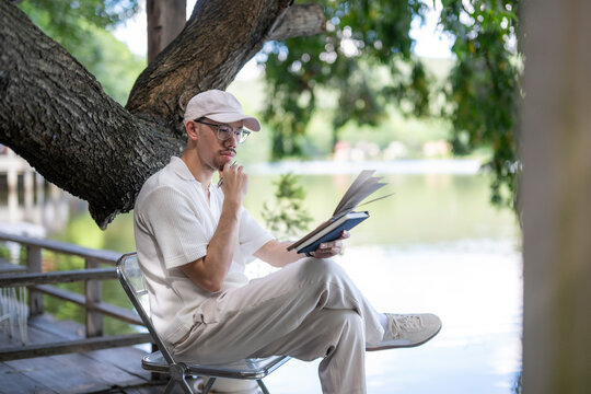 man is sitting on a chair by a lake, reading a book