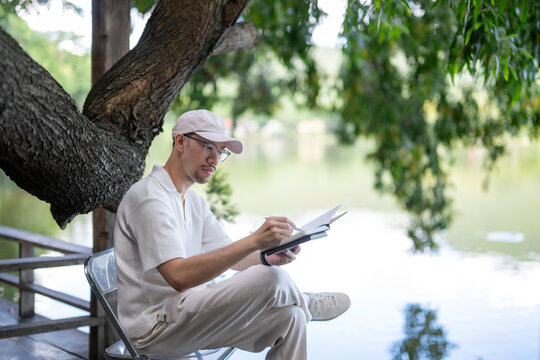 man is sitting on a bench by a lake, writing in a notebook