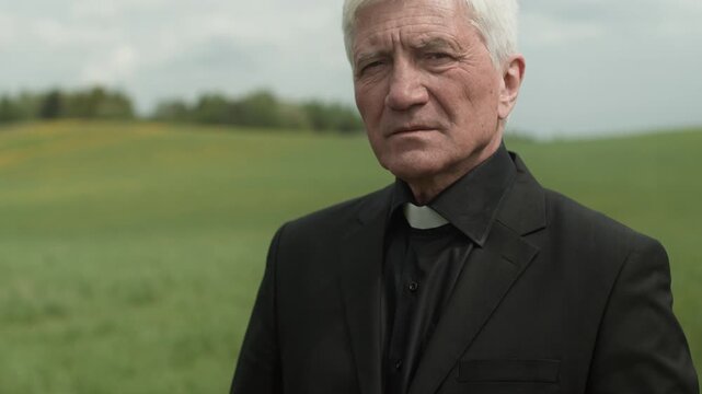 Tilt up portrait of senior Caucasian priest in black soutane holding rosary beads and looking at camera at outdoor funeral or memorial service
