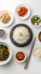 Top-down studio image of a Hanjeongsik Korean meal set with rice in a stone bowl, surrounded by colorful banchan on a white background. Includes kimchi, japchae, grilled fish, tofu