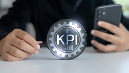 Business hand holding magnifying glass with KPI icon and gear symbol, showing key performance indicators, mobile analysis, strategic monitoring, and data tracking in modern business.