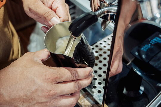 Close-up of barista steaming milk using a metal frothing pitcher and espresso machine steam wand for a hot coffee drink.