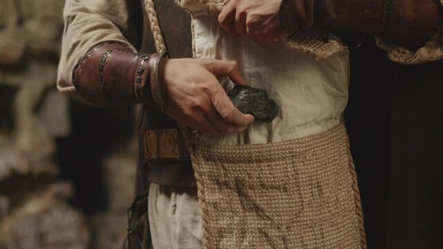 A person in historical or fantasy costume with leather armor puts a piece of rock or coal into a rustic bag. Medieval reenactment, LARP, or fantasy adventure concept.