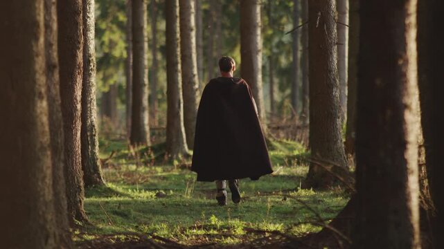 Cinematic shot of a historical or fantasy character in a dark cloak walking into a magical forest. Epic adventure, journey, or mystery concept for film and storytelling.