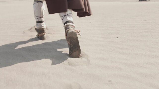 Cinematic slow motion shot following the feet of a robed character walking through a vast, sandy desert. A concept of a long, difficult journey, adventure, or exploration.