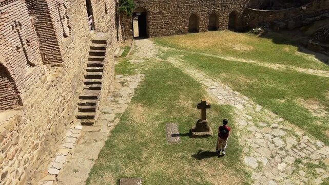 Downward shot captures traveler snapping photo at cross monument, with weathered stone walls and golden grasslands stretching across. Ideal for pilgrimage or heritage content.