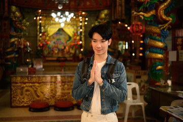 A respectful Asian traveler stands with hands pressed together inside a vibrant Chinese temple,...