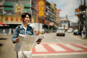 A smiling Asian male traveler stands at a crosswalk in Chiangmai, holding a manual map while...