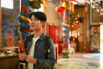 A Young Asian male traveler holding incense in a beautifully decorated Chinese temple, ready to...