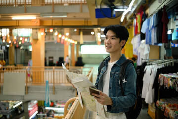 A young Asian tourist in a denim jacket stands with a paper map, planning his route inside a local...