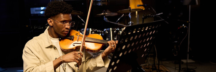 Young adult Black man playing violin while reading sheet music in music studio, focused expression, musical instruments visible in background, engaging in professional rehearsal