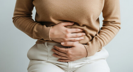 Close-up of woman's hands holding stomach area, wearing beige sweater and white pants, depicting discomfort or pain