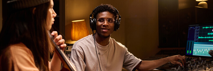 Young Black man wearing headphones operating audio mixing console while sitting in recording studio...