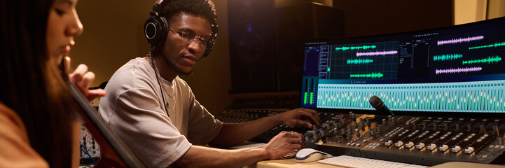Black young adult man wearing headphones operating audio mixing console in professional recording...