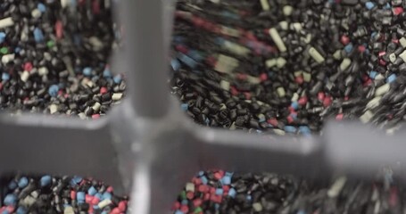 Pieces of multi-colored plastic are mixed in an industrial mixer before being disposed or reused. Close up handheld shot, real time speed, artificial indoors lighting.