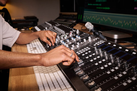 Young adult Black man adjusting audio mixing console in professional recording studio, hands operating multiple sliders and dials near computer keyboard and microphone