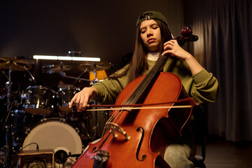 Latin girl playing cello in music studio, focused expression, long hair visible under backward cap,...