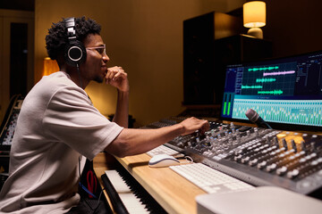 Black young adult man wearing headphones operating audio mixing console in professional recording...