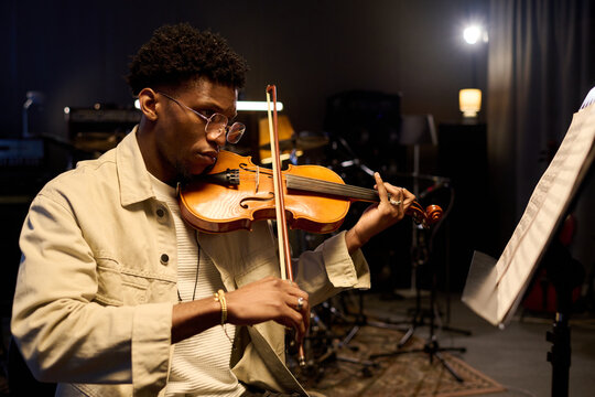 Young adult Black man playing violin in music studio, focusing on sheet music while performing, sitting on chair with musical instruments and equipment visible in background