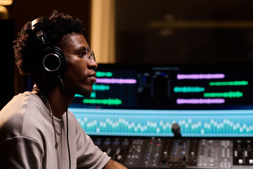 Young adult Black man wearing headphones working at audio mixing console in professional recording...