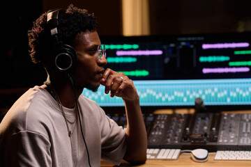 Young adult Black man wearing headphones sitting at audio mixing console working in professional...