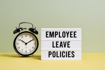 Classic black alarm clock with white face and traditional bells next to illuminated employee leave policies sign on yellow background.
