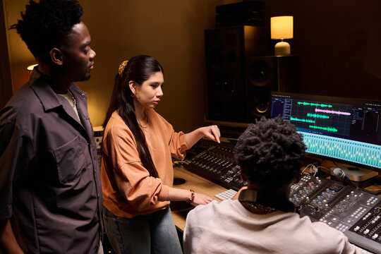 Young adult Black man, young adult Hispanic woman, and young adult Black man collaborating in professional recording studio, operating audio mixing console and computer monitors