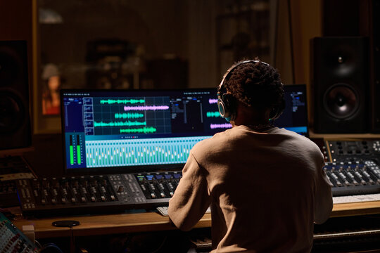 Black young adult man working at audio mixing console wearing headphones, editing music tracks on dual computer monitors in professional recording studio, focused on production - Powered by Adobe
