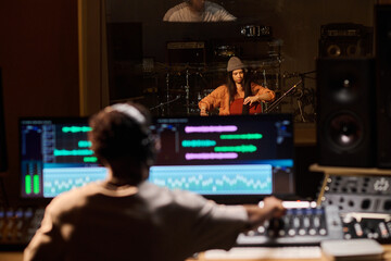Young adult Latin woman playing cello in recording studio while Black man operating audio mixing...