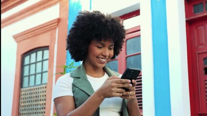 Young Black woman smiling while texting on her smartphone in a colorful colonial street. A moment of digital connection, joy and modern lifestyle. - Powered by Adobe